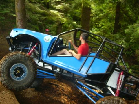 1990 Ford Bronco - A blue tube-chassis rock buggy climbs a dirt hill on a wooded trail, driven by a man in a red shirt with no doors or roof panels. It runs a…