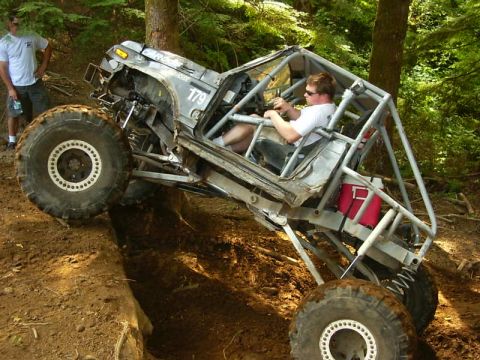 1990 Ford Bronco - A gray tube-frame rock crawler numbered 179 flexes over a dirt ledge on a wooded trail, front end lifted with a driver in a white shirt and…