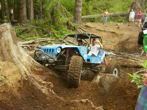 1990 Ford Bronco - A light blue Jeep-bodied buggy numbered 198 with a full black roll cage climbs out of a dirt ditch on a forest trail, driver and passenger…