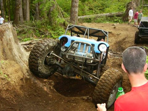 1990 Ford Bronco - A custom-built off-road buggy navigates a steep incline in a wooded area, with another vehicle visible in the background.