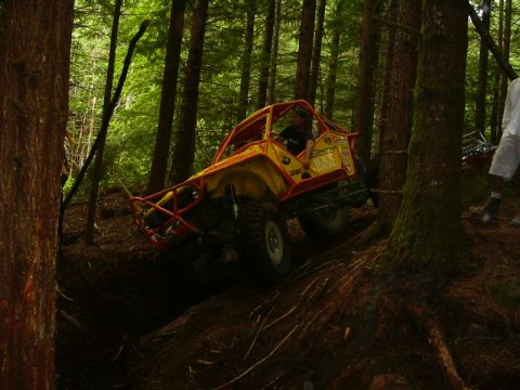 1990 Ford Bronco - A yellow rock crawler with an orange tube cage, tube front bumper and BFGoodrich and sponsor decals climbs out of a rutted dirt gully in a…