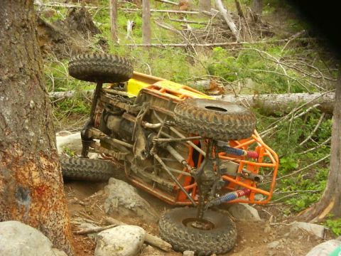 1990 Ford Bronco - An overturned off-road buggy lies on its side amidst trees and debris on a rough trail.