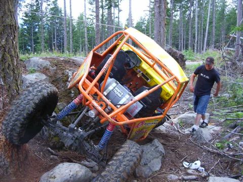 1990 Ford Bronco - An off-road buggy has tipped onto its side amidst rocks and trees, with a man observing the scene.