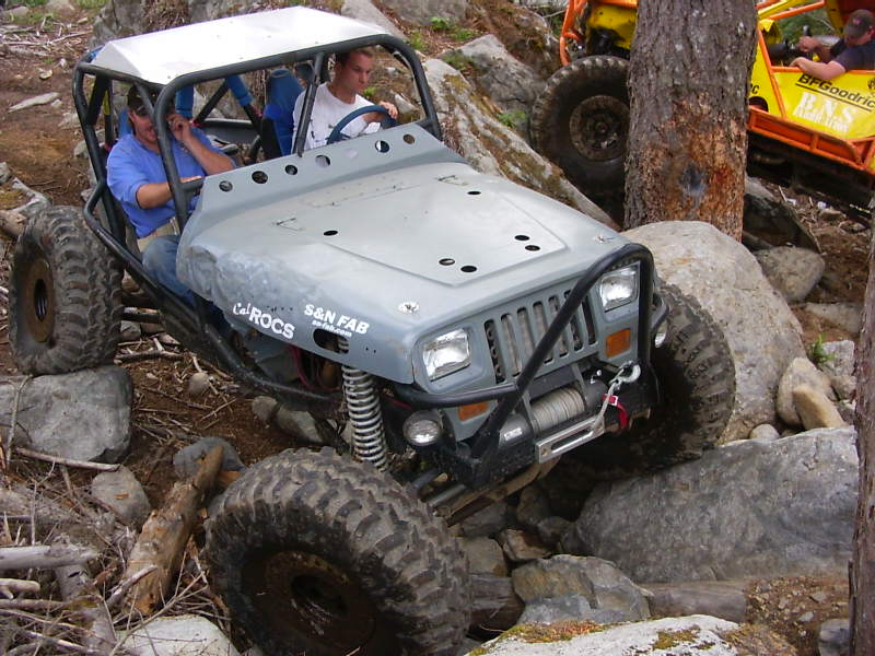 Gray custom off-road vehicle with roll cage navigating rocks. Text on side reads 'ROCS S&N FAB'