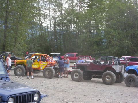 1990 Ford Bronco - A gravel staging pit at an off-road event, with a yellow tube-framed rock buggy on sponsor-decaled bodywork and a maroon doorless Jeep on o…
