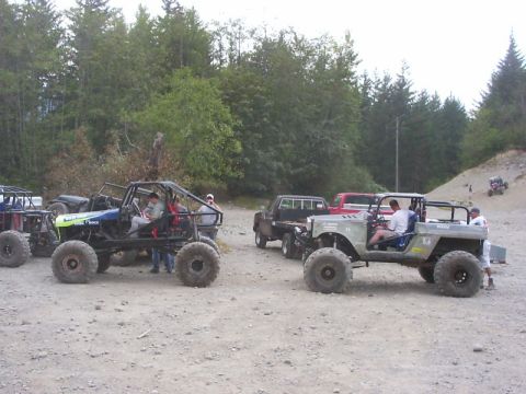 1990 Ford Bronco - A group of off-road vehicles are gathered in a wooded area, with some people standing around them.