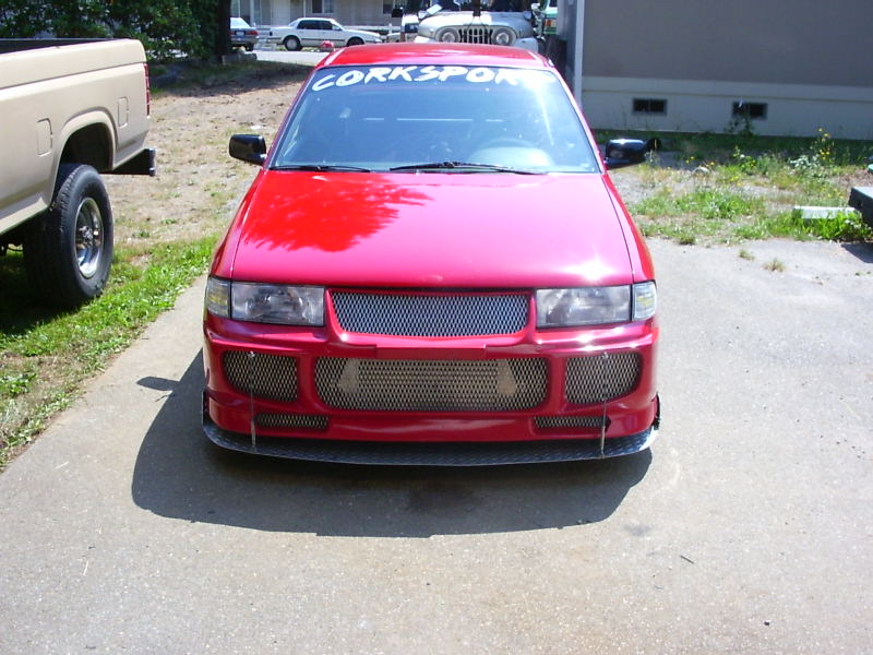 Red sedan with custom front bumper and grille, parked next to a tan pickup truck. "CORKSPORT" banner on windshield