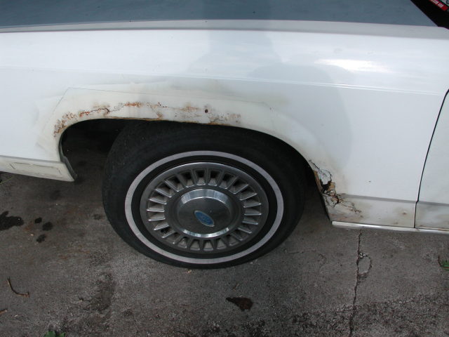 White sedan fender with rust around wheel well. Has a Ford hubcap