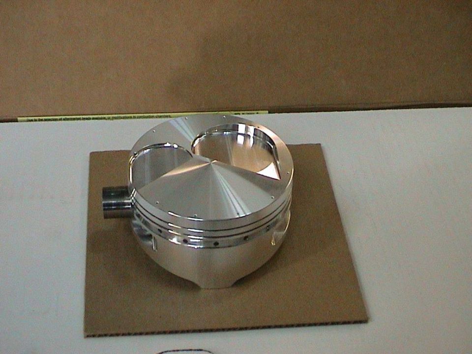 Polished aluminum piston with three valve reliefs and two wrist pin clips, resting on cardboard