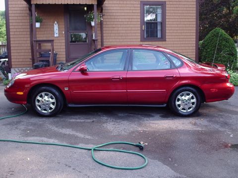 1998 Ford Taurus - A red 1998 Ford Taurus sedan is parked in front of a house, with a garden hose on the ground.