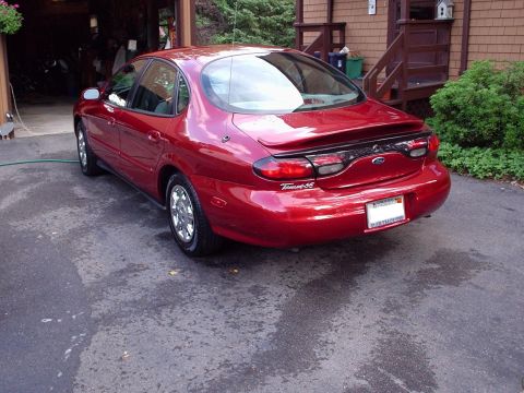 1998 Ford Taurus - A red 1998 Ford Taurus sedan is parked on a wet driveway, with a house and garage in the background.