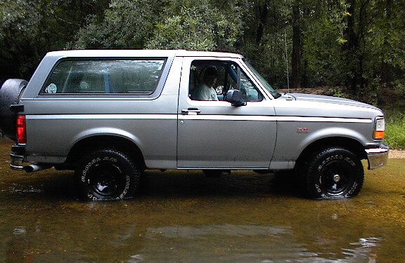 Silver Ford Bronco SUV side view, black wheels with off-road tires, sitting in shallow water in the woods