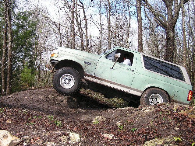 Green Ford Bronco flexing on a dirt mound with front wheel lifted on a wooded trail