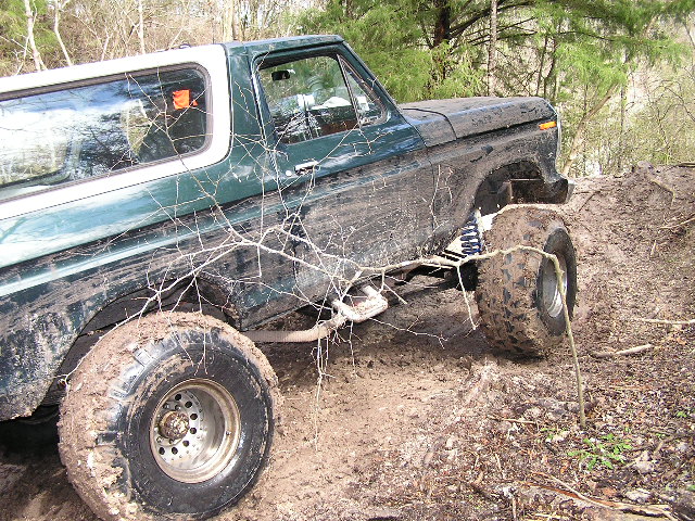 Dark green and white Ford Bronco climbing a muddy off-camber bank, front tire stuffed