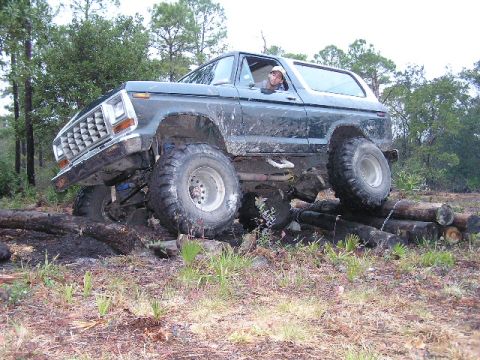 1994 Ford Bronco - A dark green Ford Bronco is shown off-road, navigating over a stack of logs.