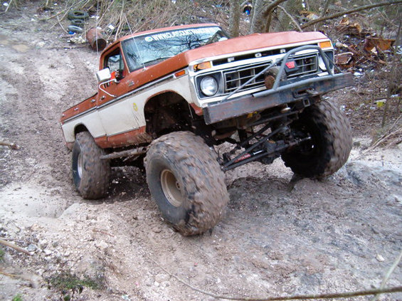 Lifted orange and white 1970s Ford pickup on huge mud tires flexing on a rutted trail