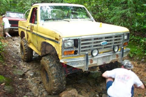 1985 Ford Bronco - Three-quarter front view of a battered yellow lifted Ford Bronco with a white top, red interior roll bar, dented fenders and surface rust o…
