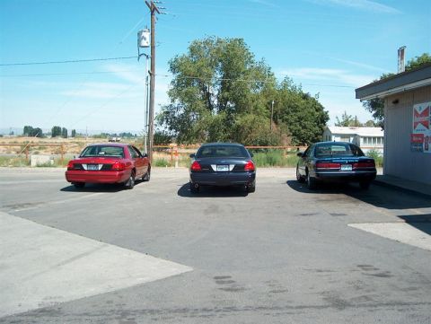 2001 Ford Crown Victoria - Three sedans are parked in a lot on a sunny day, with a building to the right and trees in the background.