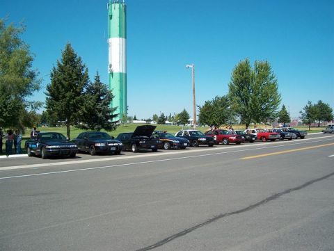 2001 Ford Crown Victoria - A line of cars, including a black police car, are parked near a water tower under a clear blue sky.
