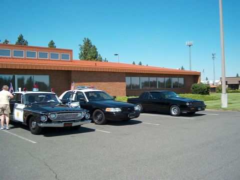 2001 Ford Crown Victoria - Three police cars, including a vintage model and two modern sedans, are parked in a lot outside a brick building.