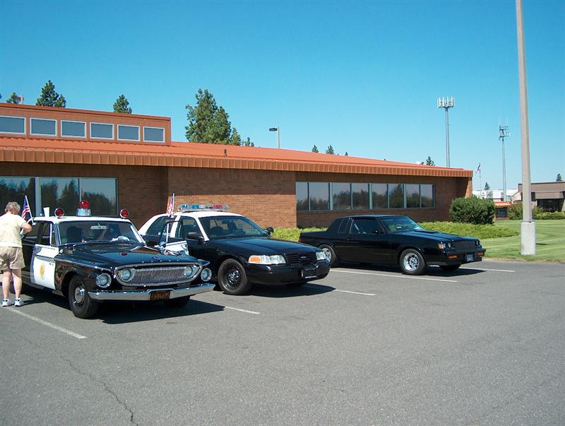 Three police cars parked in a lot: a vintage black and white sedan, a black Ford Crown Victoria, and a black coupe