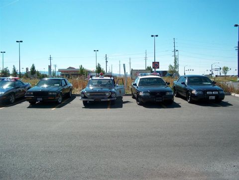 2001 Ford Crown Victoria - A lineup of police vehicles, including a vintage model and several modern sedans, are parked in an outdoor lot under a clear blue sky.