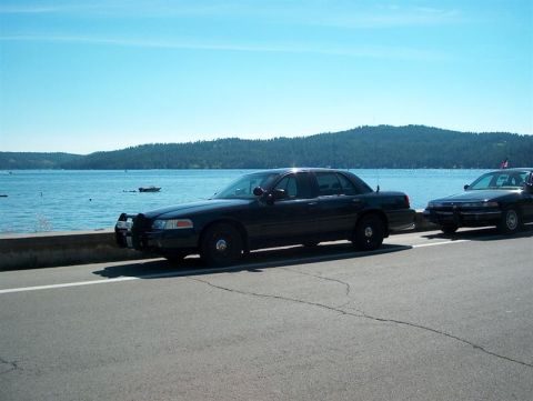 2001 Ford Crown Victoria - Two black Ford Crown Victorias are parked by a lake with hills in the background.