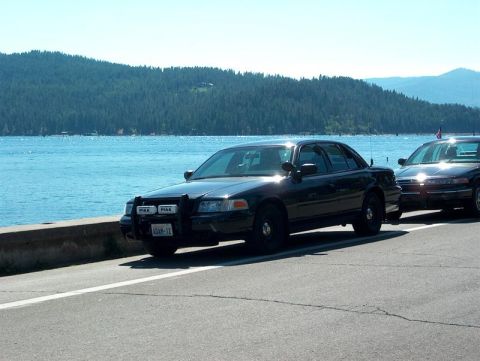 2001 Ford Crown Victoria - Two Ford Crown Victoria police cars are parked along a scenic waterfront road with mountains in the background.