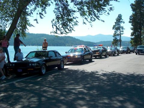2001 Ford Crown Victoria - A black coupe is parked near a body of water, with several police vehicles lined up behind it on the road.