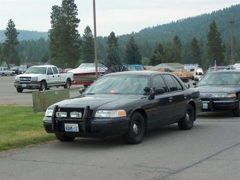 2001 Ford Crown Victoria - A black police car with a push bar is parked on asphalt, with other vehicles and a boat trailer in the background.
