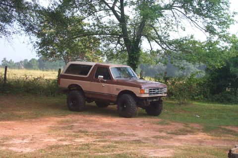 1987 Ford Bronco - This lifted 1987 Ford Bronco is parked outdoors on a dirt area with trees and a field in the background.