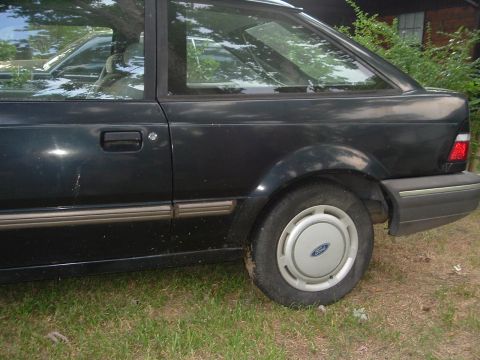 1989 Ford Escort - This dark sedan, possibly from the late 1980s, is parked on a grassy area next to a building.