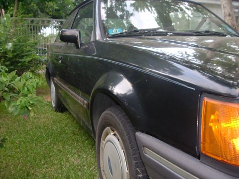 1989 Ford Escort - This car is parked on a grassy area next to some bushes and a fence.