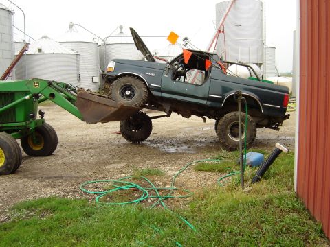 1992 Ford Bronco - A tractor is lifting a 1992 Ford Bronco by its front bumper with the hood open.