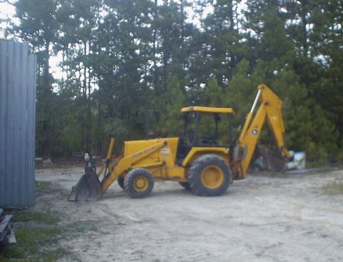1989 Ford Bronco - A yellow John Deere backhoe loader is parked on a dirt lot next to a metal building and trees.