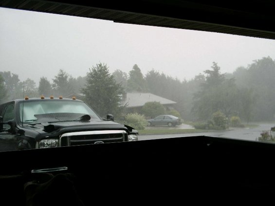 View from a garage of a black Ford Super Duty in heavy rain, gray sedan and houses beyond