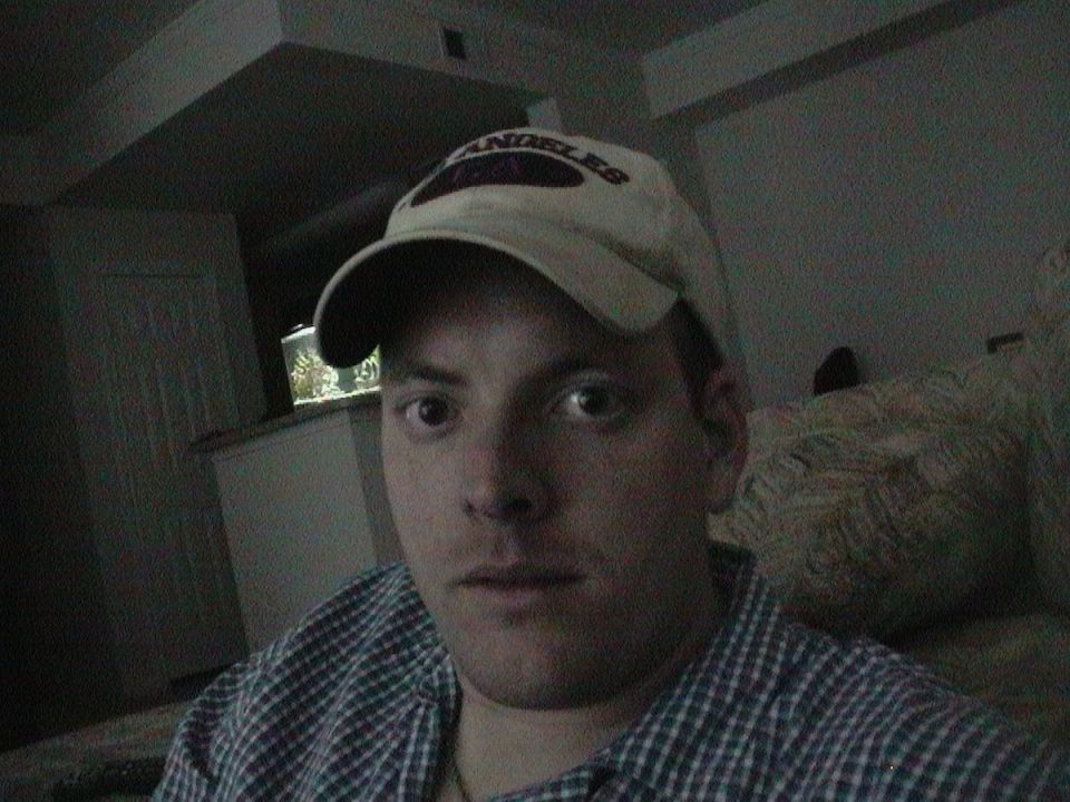 Young man in a white Los Angeles ball cap and plaid shirt taking a selfie in a dim room