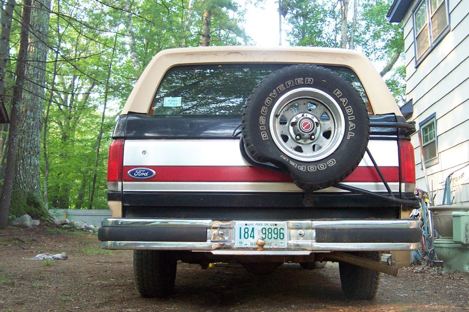 Rear of tan and black Ford Bronco with Cooper spare tire on tailgate carrier, NH plate, parked by a house
