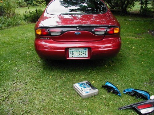 Red 1997 Ford Taurus rear with detached tail lights and a toolbox on the grass