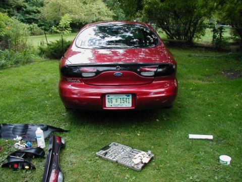1997 Ford Taurus - A red 1997 Ford Taurus is parked on grass with its rear facing the camera, surrounded by car parts and tools.