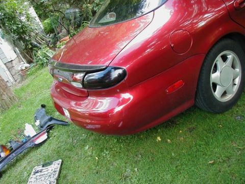 1997 Ford Taurus - The rear section of a red sedan is shown with aftermarket blacked-out tail lights and a removed bumper trim piece on the grass.