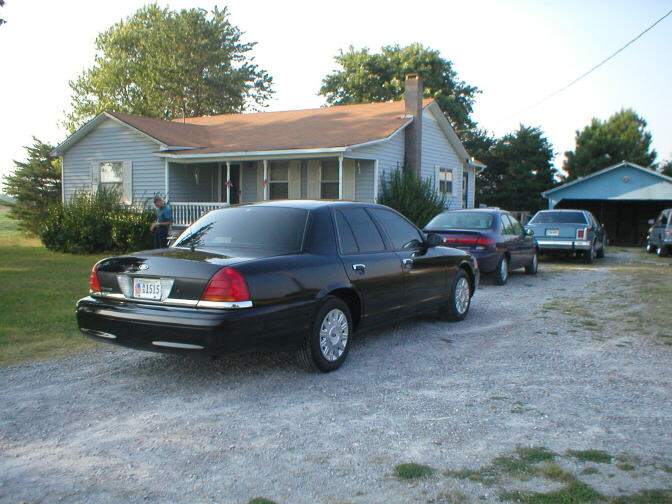 Black sedan parked in front of a house, with other cars behind it. License plate is visible