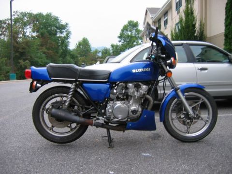 1985 Ford Crown Victoria LTD - A blue Suzuki motorcycle is parked on asphalt, with a silver car and a building behind it.