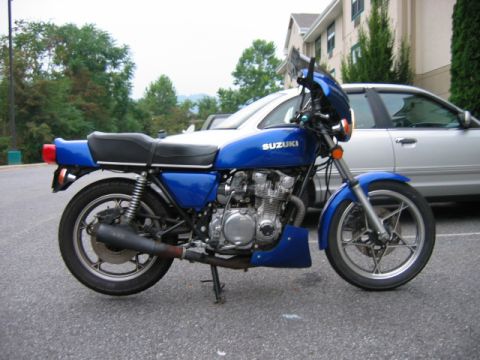 1985 Ford Crown Victoria LTD - A blue Suzuki motorcycle is parked on asphalt, with a silver sedan in the background.