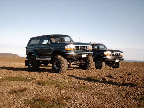 1996 Ford Bronco - Two lifted Ford trucks, a green Bronco and a black F-Series, are parked on a rocky, arid landscape under a clear blue sky.