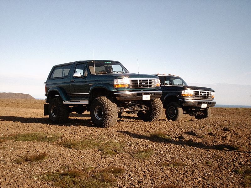 Green lifted Ford Bronco and blue Ford pickup on gravel under clear sky