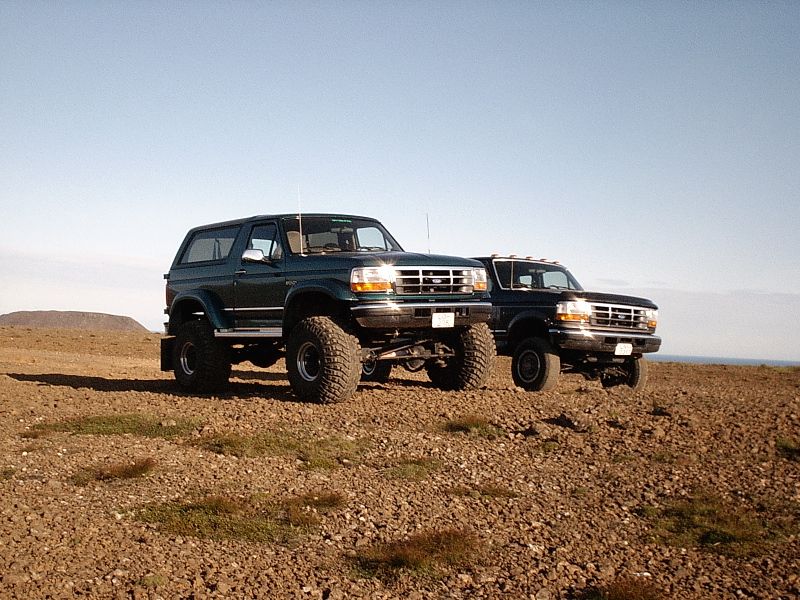 Lifted teal Ford Bronco on large tires beside a lifted Ford pickup on gravel terrain.