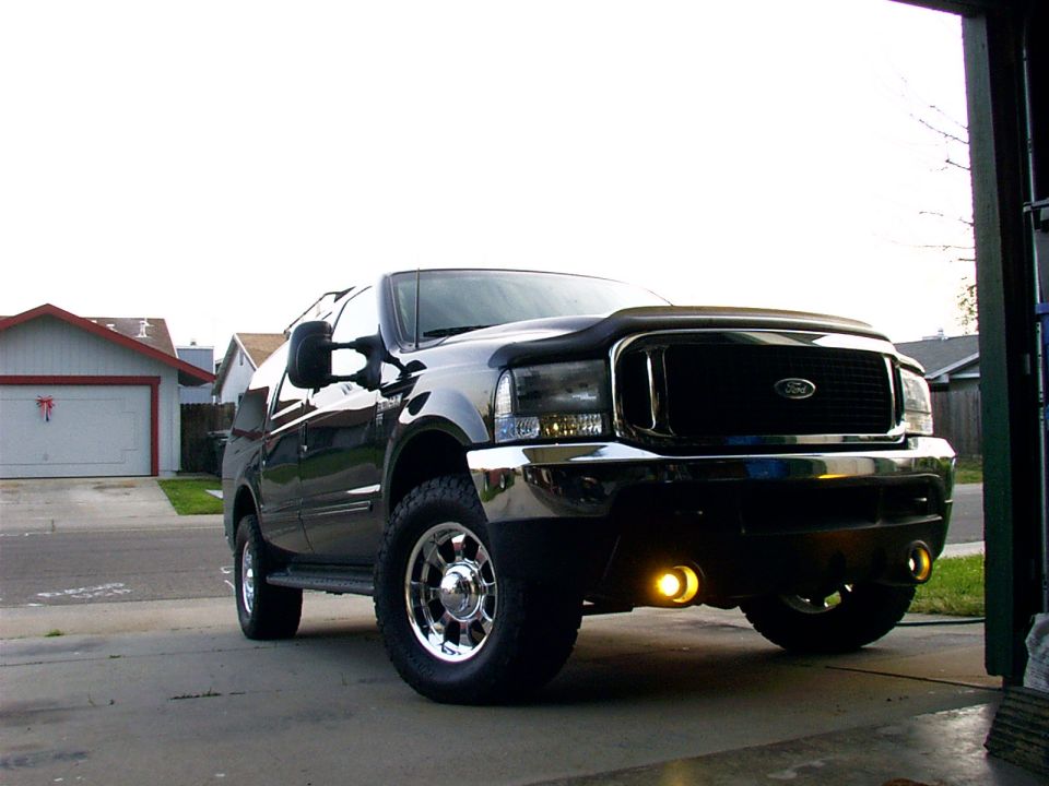 Black Ford Excursion SUV with chrome grille, wheels and lit amber fog lights, viewed from low front angle in a driveway