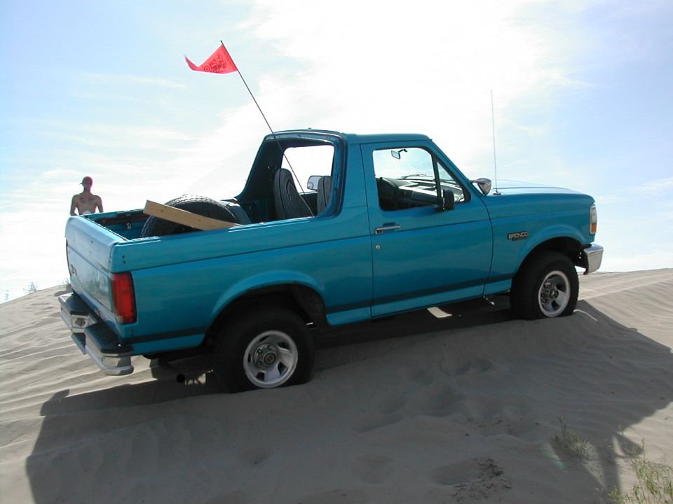 Teal Ford Bronco with a red flag on a sand dune, driver's side visible