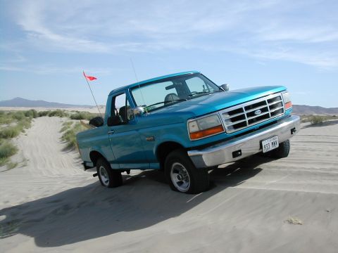 1994 Ford Bronco - A blue pickup truck navigates a sandy terrain under a clear sky.